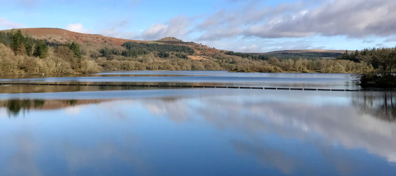Clouds are reflected in the still water of the pond on a bright day. In the background is the moors and trees surrounding the waters edge. 