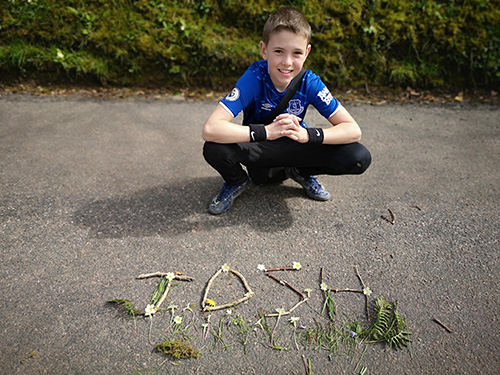 Boy smiling next to sticks and greenery spelling out 'Josh