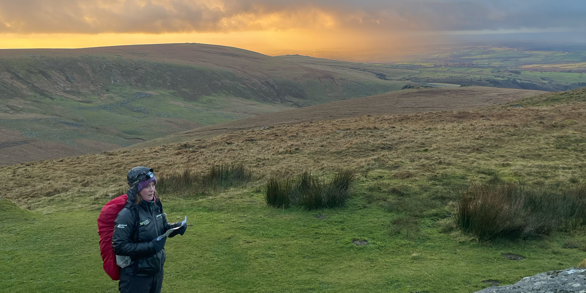 Female ranger map reading. Behind the moor stretches out as the sun sets in the sky. 