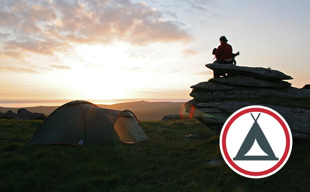 Small green tent and person sat on a tor with dog at sunset